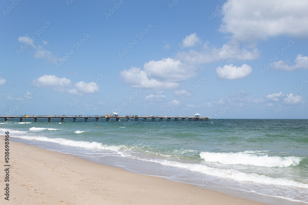 Beach on a sunny day with a calm sea, a sky with few clouds and a pier on the horizon line