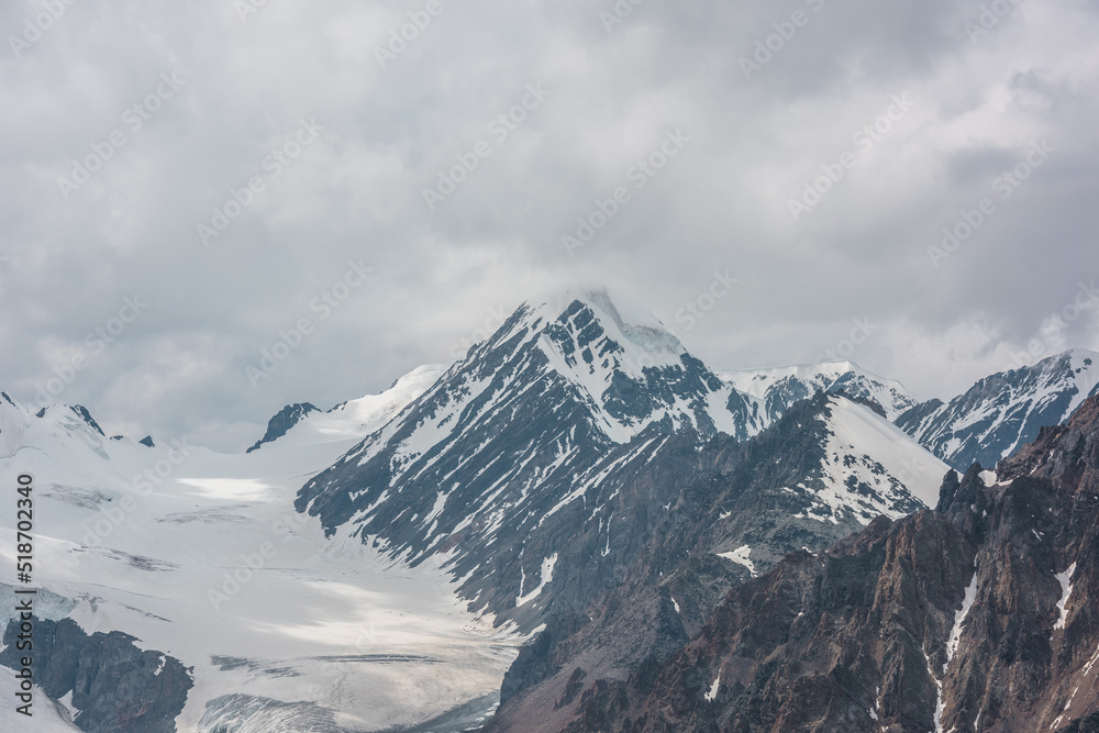 Obraz premium Atmospheric landscape with sharp rocks and high snowy mountain top in rainy low clouds at overcast. Dramatic gloomy scenery with large snow mountains and glacier in gray cloudy sky at rainy weather.