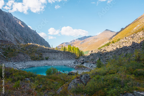 Pure turquoise alpine lake among lush autumn vegetation with view to large mountains in sunlight. Beautiful glacial lake against sunlit pyramid shaped mountain. Vivid autumn colors in high mountains.
