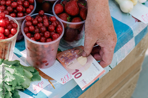 Selling berries at the farmers' market. Cherries and strawberries in plastic glasses. Elderly woman's hand holding a 10 euro banknote. Organic food. Trade concept. photo