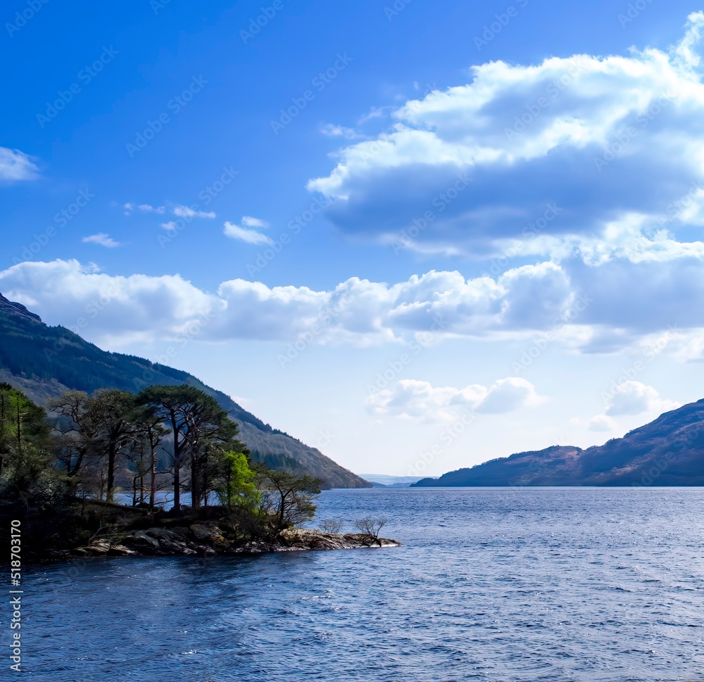 Beautiful Loch Lomond, Scottish Highlands. Calm
water and blue sky with clouds. Bit of land with trees
jutting out into the lake. Peaceful, serene scenic view.