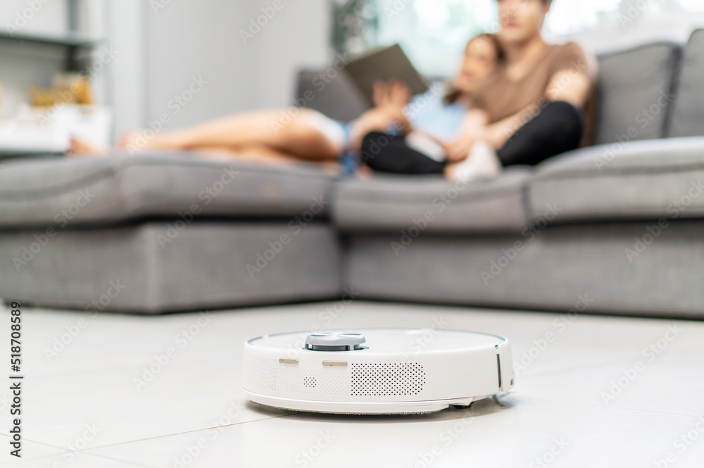 Fototapeta premium Young couple resting on the couch while robotic vacuum cleaner doing its work at home. Household robots, Smart home concept