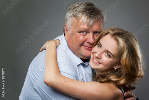 Dad and teenage daughter laugh and hug. A fat man in a shirt and a beautiful girl in a blue dress on a dark gray background. Love and tenderness. Close-up.