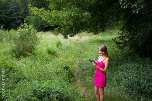 A slender young freelancer girl with long blond hair, in a pink dress, stands in the forest on a green lawn and remotely works and studies on a tablet.