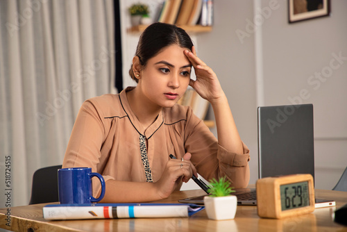 A young modern Indian Asian corporate woman or female entrepreneur is working on a project or challenging task using a laptop thinking, worried, and stressed sitting in a start up business office. photo