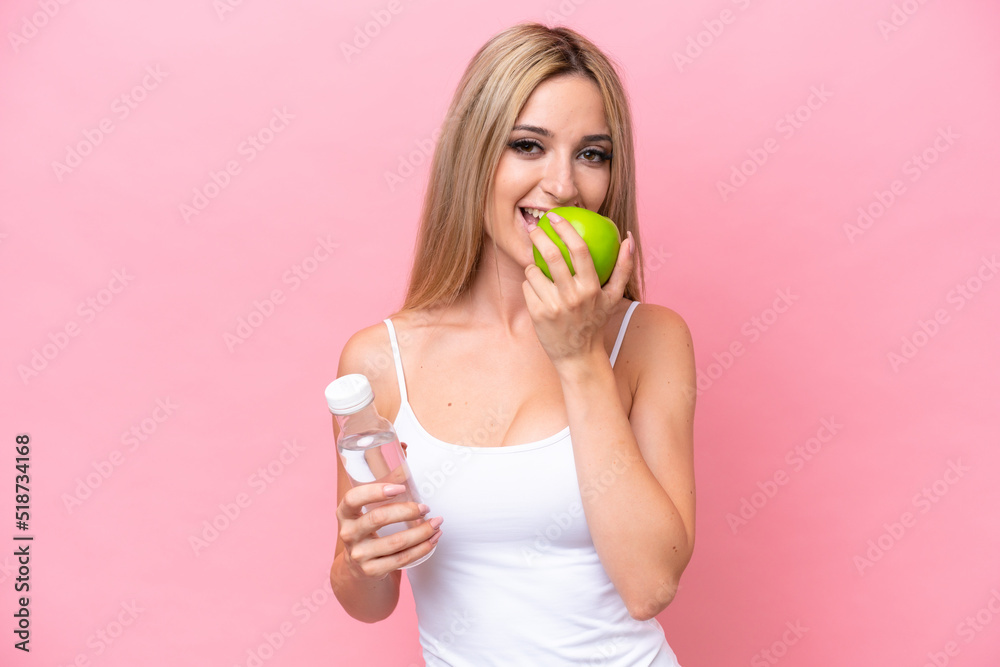 Pretty blonde woman isolated on pink background with a bottle of water and eating an apple Pretty blonde woman isolated on pink background with a bottle of water and eating an apple