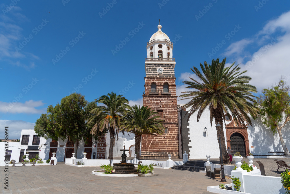 Plaza del pueblo costa Teguise en Lanzarote, con un gran campanario de ladrillo al fondo, rodeado por palmeras en un día soleado con el cielo azul despejado en las islas Canarias.