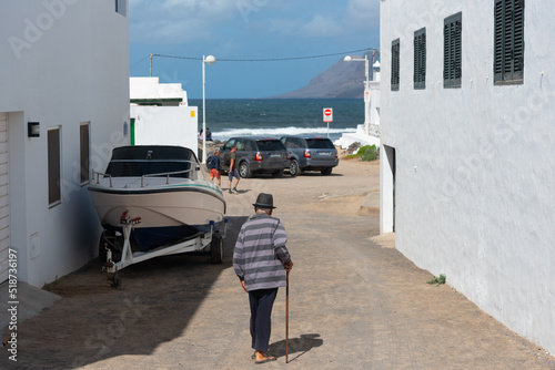 Hombre mayor con sombrero, camiseta de rayas y bastón, de espaldas paseando por las calles con el mar al fondo en Lanzarote durante un día soleado con el cielo azul  despejado, Islas Canarias photo
