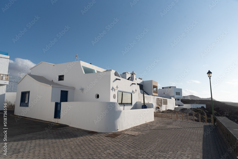 Pueblo La Santa en Lanzarote, vista de sus  calles y casas con la tradicional arquitectura de las Islas canarias en color blanco y de líneas simples, al fondo grandes montañas volcánicas.