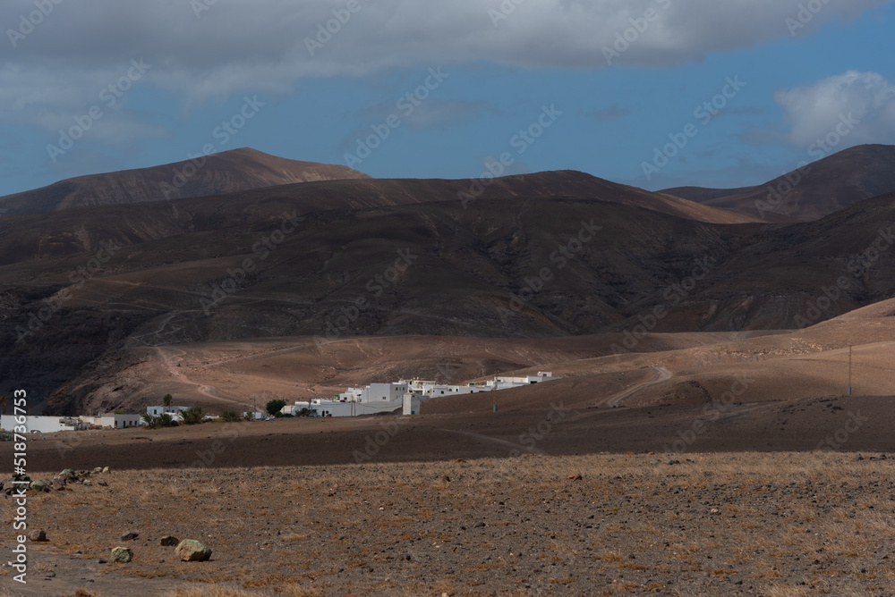 Vista panorámica de un impresionante paisaje volcánico y desértico con un pueblito de casas blancas y pequeñas montañas durante un día soleado con el cielo azul y nubes en Lanzarote Islas Canarias.