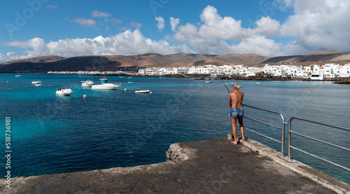 Fototapeta Naklejka Na Ścianę i Meble -  Mujer rubia pescando frente a Punta Mujeres  con pequeñas casas blancas típicas de Lanzarote en la costa junto al mar turquesa con barcos y volcanes inactivos detrás un día soleado en Islas Canarias
