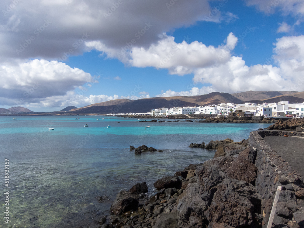 Pueblo de Punta Mujeres con casas tradicionales de color blanco en la isla de Lanzarote en Islas Canarias rodeado por un mar turquesa y volcanes inactivos al fondo durante un día soleado y despejado.
