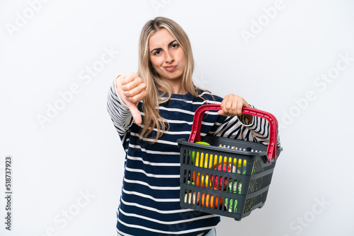 Young caucasian woman holding a shopping basket full of food isolated on white background showing thumb down with negative expression