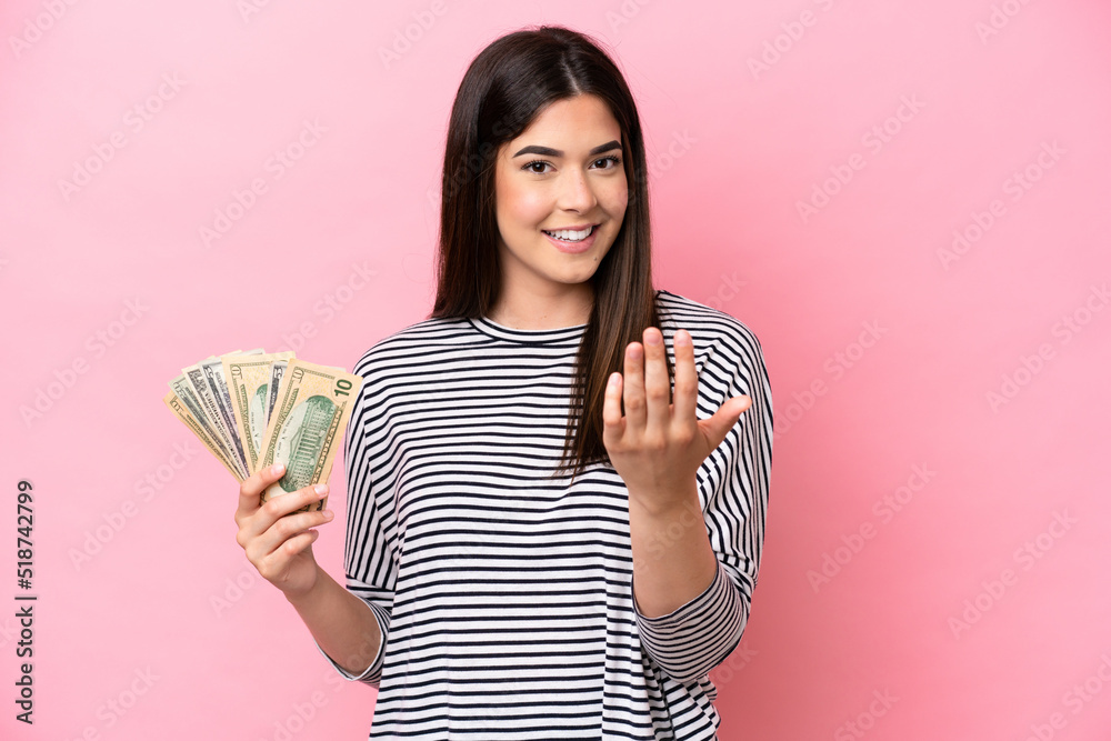 Young Brazilian woman taking a lot of money isolated on pink background inviting to come with hand. Happy that you came Young Brazilian woman taking a lot of money isolated on pink background inviting to come with hand. Happy that you came
