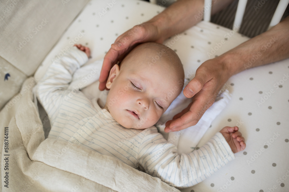 Naklejka premium male hands carefully hold the head of a sleeping baby, a small child sleeps in his crib, a child's daytime sleep