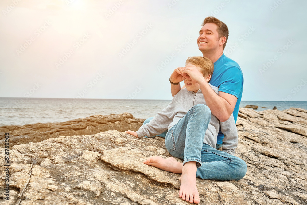 Happy family, father and son bonding, sitting on stone by the sea looking at view enjoying summer vacation. Togetherness Friendly concept
