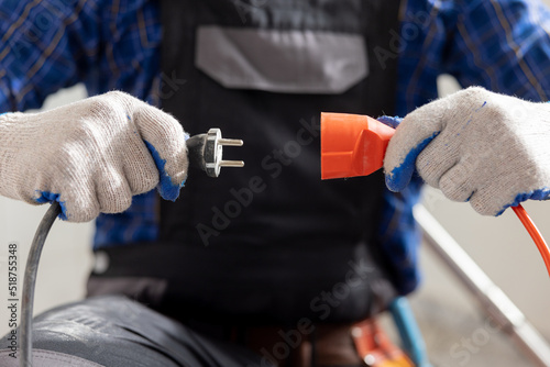 Hands of a man wearing work gloves hold an extension cord and cable connection of two plugs plugging the equipment into electricity.