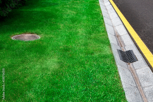 gray gutter of a stormwater system on the side of asphalt road with yellow markings and manhole in green trimmed lawn, concrete drainage ditch with iron grate on sunny dry weather, nobody. photo