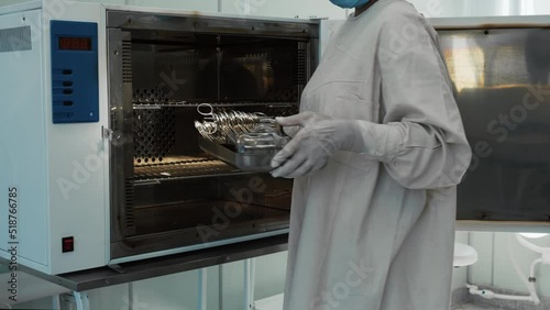 Female nurse in white protective suit taking clean sterilized surgical tools and instruments from medical autoclave steam sterilizer machine, healthcare worker in hospital operating room, slow motion.