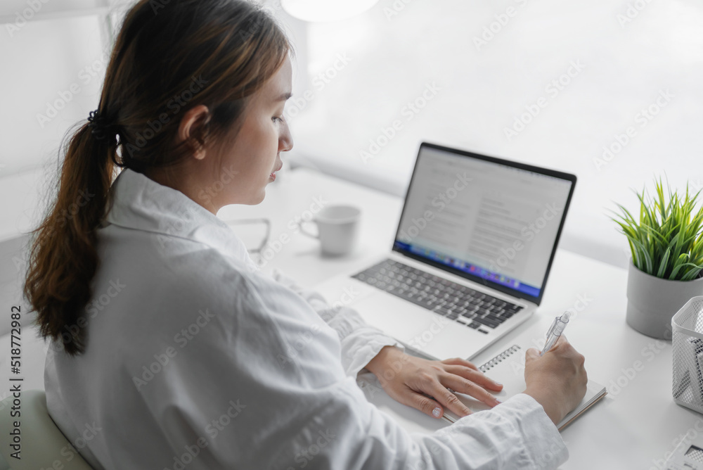 Asian businesswoman taking notes in notebook while using laptop at home office Asian businesswoman taking notes in notebook while using laptop at home office