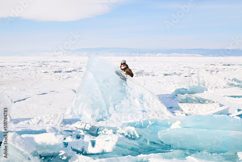 A young woman stands behind a huge block of ice. Piles of blue ice fragments, hummocks covered with snow. Frozen Lake Baikal on a sunny winter day.