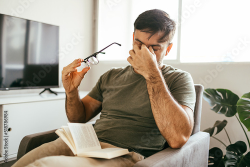 Tired man takes off his glasses and rubs his eyes while reading a book at home. photo