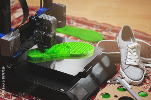 3D-printer makes bright green shoe sole from flexible plastic filament in comfy home surrounding. direct drive extruder, metal print bed, grey sneaker shoes and filament spool.  selective focus photo