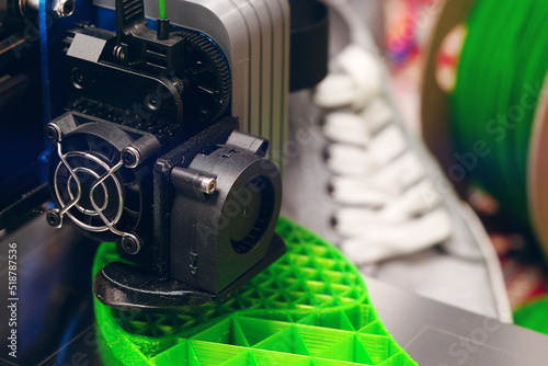 print head of 3D-printer makes a shoe sole with distinct inner structure from bright green plastic filament. closeup view with grey sneaker shoes and filament spool in background. selective focus photo