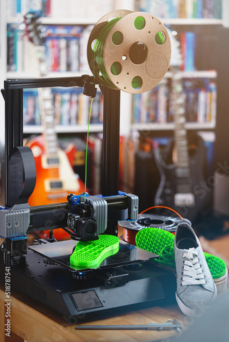 3D-printer produces a custom bright green shoe sole in living room surrounding. total view with grey sneaker shoes and filament spools. Guitars in background. upright orientation. selective focus.