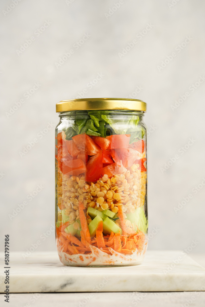 Trendy Fresh detox summer salad layered in a glass mason jar: yoghurt dressing, carrots, cucumber, yellow lentils, red bell pepper, green leaves and herbs, long silver spoon, on white background Trendy Fresh detox summer salad layered in a glass mason jar: yoghurt dressing, carrots, cucumber, yellow lentils, red bell pepper, green leaves and herbs, long silver spoon, on white background