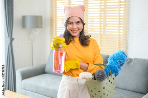 Young happy woman wearing yellow gloves  and holding a basket of cleaning supplies in living room.