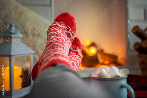 Cozy winter evening at home, feet in warm woolen red socks by the fireplace, a woman is relaxing and with a mug of hot drink with marshmallows and enjoying the comfort. Christmas holiday concept.
