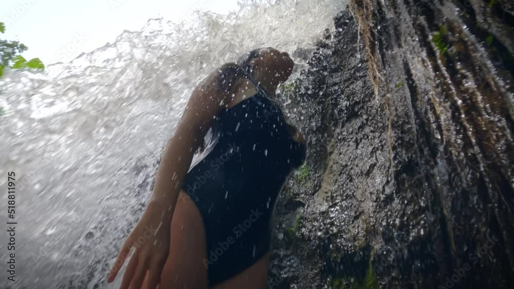 A slender girl in blue swimsuit stands under flow of a tropical waterfall waterfall. a sexy girl