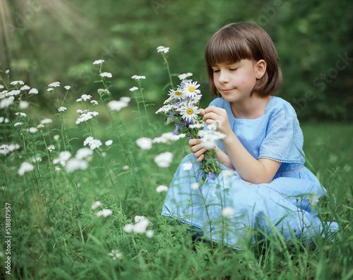 cute little girl in a blue dress with a bouquet of daisies sits on a flowers meadow in the park