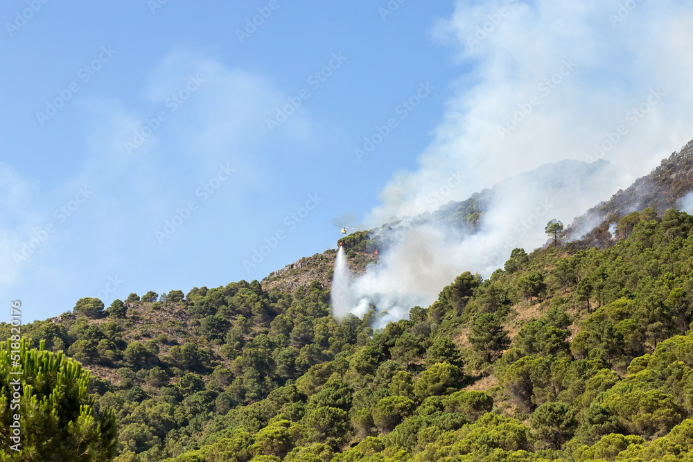Helicopter in flight putting out the fire near Pinos de Alhaurín. Dropping water on the fire on the mountainside. Pine forest burning, fire active and lots of smoke. Malaga, Spain. July 2022