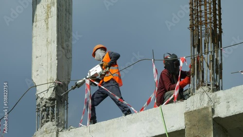 Builders work at a construction site, one with a jackhammer, the other with electric welding.
