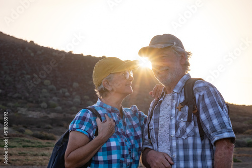 Caucasian smiling senior couple with caps and backpacks travelling in countryside landscape at sunset light - elderly man and woman enjoying freedom and healthy lifestyle