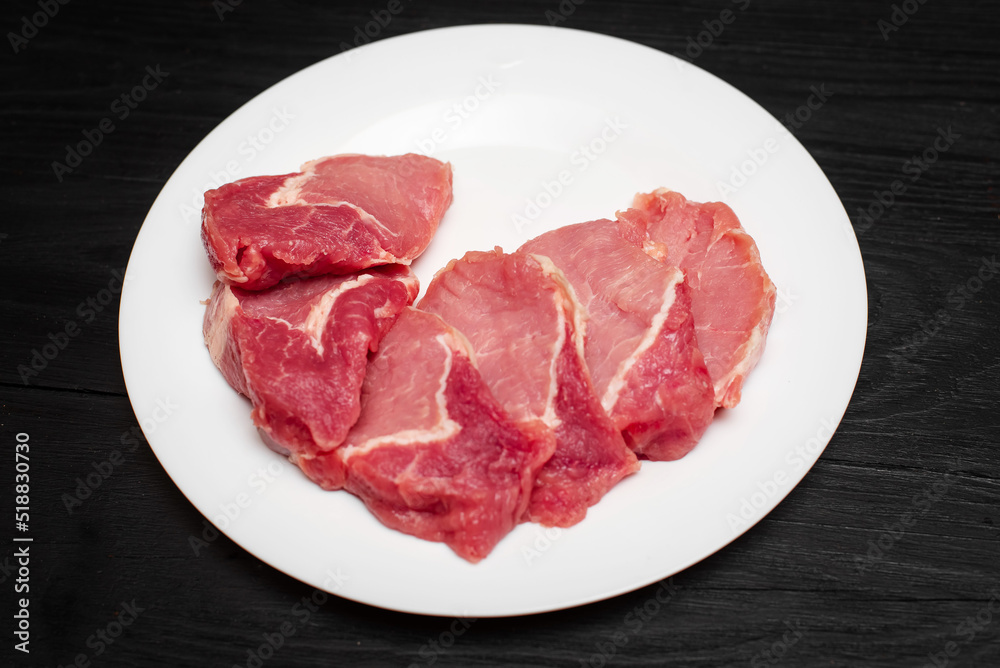 Raw pork chop on a white plate on a black wooden background. Fatty meat beaten off for cooking. Raw pork chop on a white plate on a black wooden background. Fatty meat beaten off for cooking.