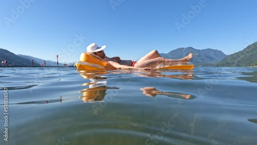 Mid adult woman lying on inflatable mattress, wearing a white straw sun hat that obscure her face while floating on the beautiful Harrison Lake surrounded with mountains in Harrison Hot Springs