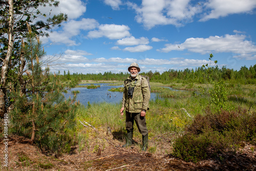 A man with a beard in camouflage clothes, with binoculars, an explorer, a tourist. Stands against the background of a forest lake, forest and blue sky with clouds.