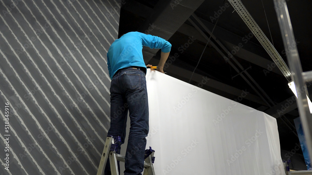 Worker builds a concert stage. Worker in blue shirt standing on stepladder, installing big stand with white, stretched cloth.
