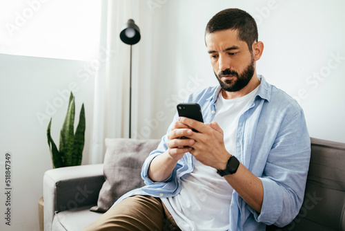 Konstfotografi Young brazilian man sitting on sofa using smartphone