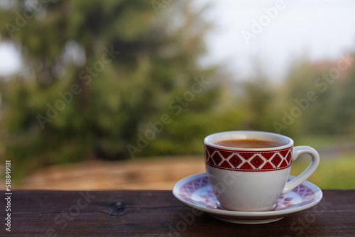 cup of goffee on wooden table. green pine tree in the background.