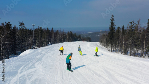 Modern ski resort in pine forest aeria, view from above. Footage. Young group of people snowboarding and skiing down the snowy slope. photo
