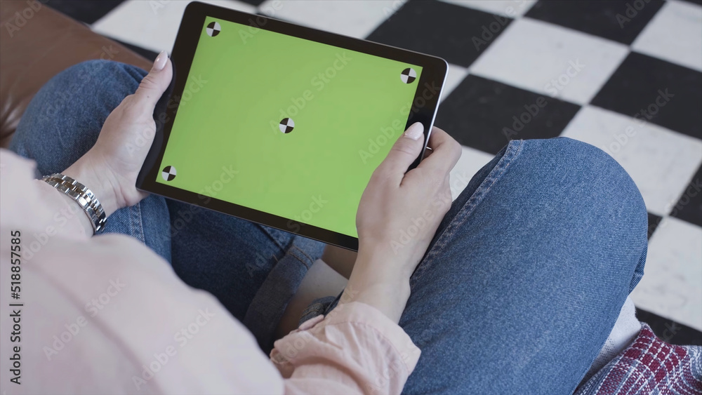 Obraz premium Young woman using black tablet device with green screen. Stock footage. Woman holding tablet, zooming pages while sitting on the couch in the living room. Chroma key.