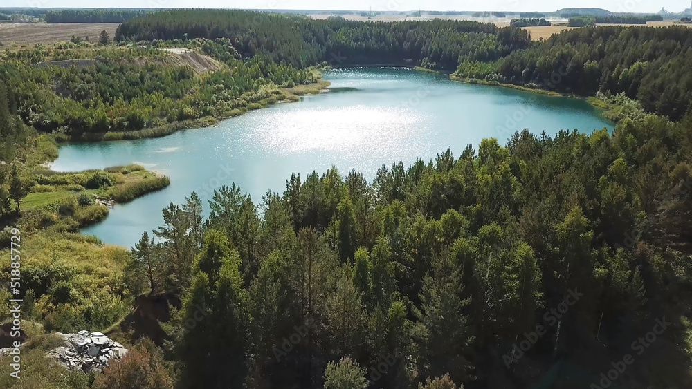 Aerial view of green forest and lake shore. Stock footage. Flying over breathtaking summer natural landscape with turquoise lake surrounded by pine tree forest.