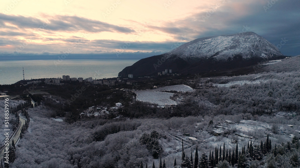Panoramic view of the embankment of the city and snow-capped high mountain in sunset. Shot. Aerial for a coastal town, winter trees and fields, high mountain covered with snow against blue sea.