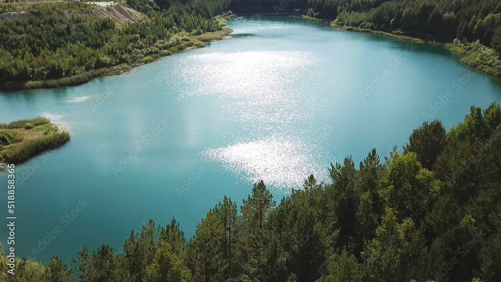 Aerial view of green forest and lake shore. Stock footage. Flying over breathtaking summer natural landscape with turquoise lake surrounded by pine tree forest.