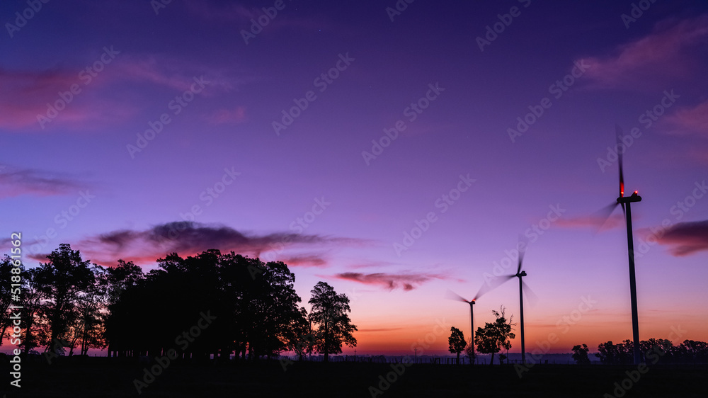 Fototapeta premium The silhouette of three modern windmills in a colorful sunset, next to a group of trees on the horizon, on the outskirts of Kiyú, San José, Uruguay