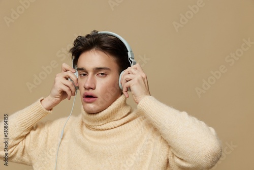 A handsome man with light white skin, with large headphones on his head with dark, short, short hair combed back, in a beige turtleneck with a high collar stands on a dark beige background.Close-up